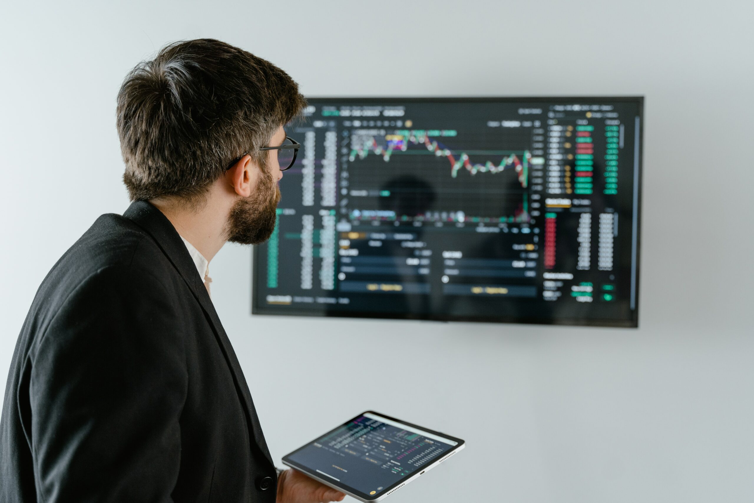 Trader mit Tablet in der Hand steht vor Bildschirm mit Charts 